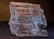 A close-up of a rough, translucent mineral specimen with visible striations and textured surfaces. The mineral displays varying shades of gray and white, with some darker inclusions. The lighting highlights the layered structure, emphasizing the natural beauty of the mineral.