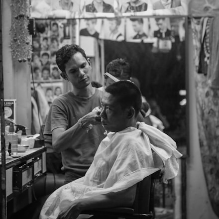 A barber is carefully trimming a customer's hair inside a small, cluttered barbershop. Various haircut styles are visible on the wall in the form of photographs. The setting has a personal and intimate ambiance, with grooming tools and products scattered around the mirror.