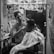 A barber is carefully trimming a customer's hair inside a small, cluttered barbershop. Various haircut styles are visible on the wall in the form of photographs. The setting has a personal and intimate ambiance, with grooming tools and products scattered around the mirror.