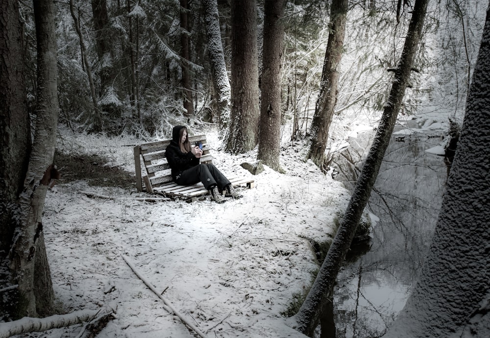 woman wearing black denim jeans sitting on bench surrounded by trees and covered with snow near body of water