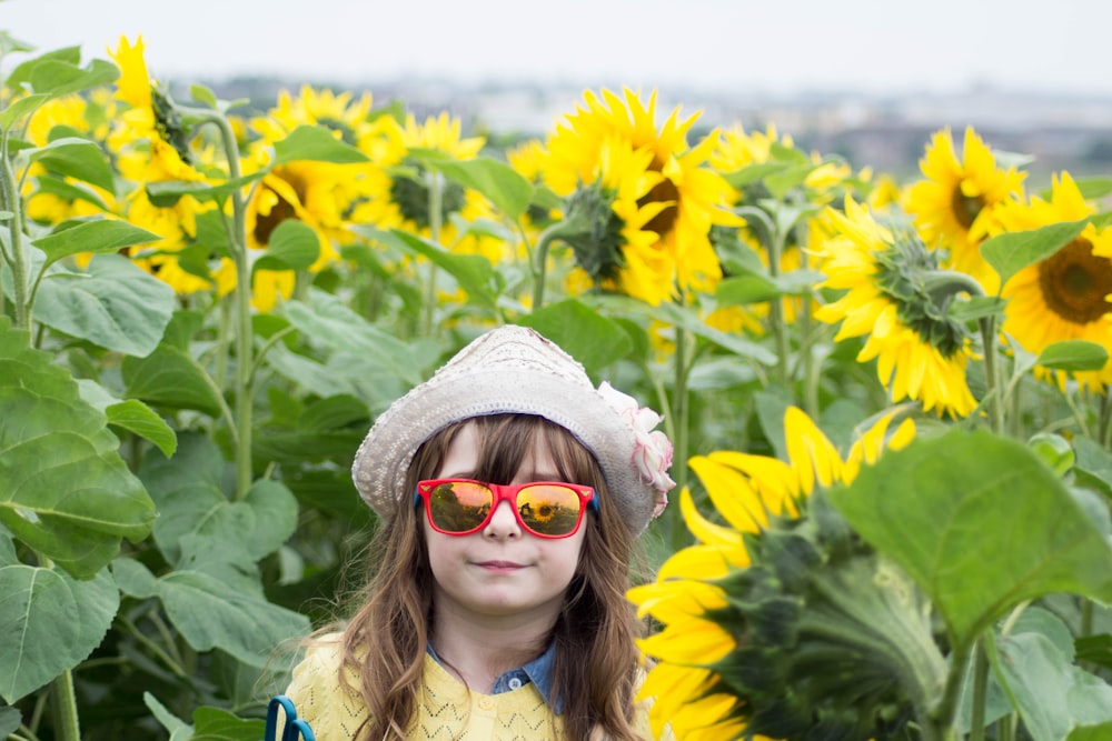 Ragazza che indossa occhiali da sole con cornice rossa e cappello bianco circondato da girasoli gialli durante il giorno