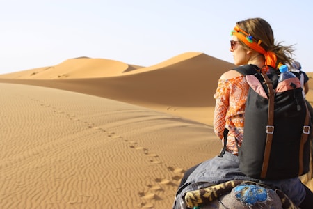 A person wearing an orange-patterned shirt, sunglasses, and an orange headband is seen riding on a camel through a vast desert landscape. The sandy dunes stretch into the distance, with visible footprints tracing a path through the sand. The person is carrying a black backpack with a water bottle attached.
