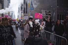A crowd of people, including adults and children, participate in a street protest in an urban setting. Many hold signs with messages advocating for women's rights and environmental issues. The scene includes a mix of genders and ages, with some individuals wearing pink hats. Tall buildings line the street as steam rises from vents.
