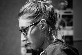 A black and white profile portrait of a person with glasses and tied-back hair. The lighting highlights the contours of their face and the details in their hair, creating a soft contrast with the dark background.