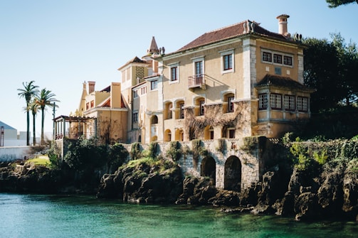 A large, old-fashioned villa with light yellow walls and red-tiled roofs is situated by the water's edge. The architecture includes elements like chimneys, wooden windows, and decorative stone arches at the base. The surrounding area features lush greenery, palm trees, and a rocky waterfront.