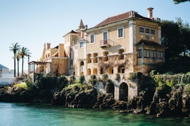 A large, old-fashioned villa with light yellow walls and red-tiled roofs is situated by the water's edge. The architecture includes elements like chimneys, wooden windows, and decorative stone arches at the base. The surrounding area features lush greenery, palm trees, and a rocky waterfront.