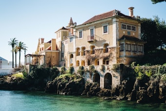 A large, old-fashioned villa with light yellow walls and red-tiled roofs is situated by the water's edge. The architecture includes elements like chimneys, wooden windows, and decorative stone arches at the base. The surrounding area features lush greenery, palm trees, and a rocky waterfront.