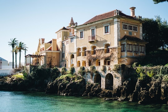 A large, old-fashioned villa with light yellow walls and red-tiled roofs is situated by the water's edge. The architecture includes elements like chimneys, wooden windows, and decorative stone arches at the base. The surrounding area features lush greenery, palm trees, and a rocky waterfront.