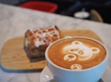 A cup of coffee with intricate latte art depicting a bear's face sits on a marble counter. In the background, a pastry glazed with icing rests on a wooden board.