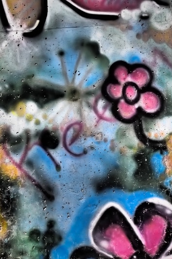 A vibrant graffiti art piece featuring a pink flower with a black outline and a portion of a butterfly with pink and black wings. The background includes blue, yellow, and green splashes of color, with some white expressive strokes and indistinct black dots. The word 'free' is partially visible in a fluid, red handwriting style.