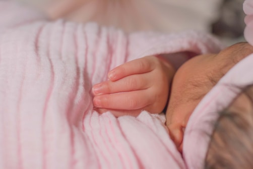 A close-up of a sleeping baby wrapped in a soft, pink blanket. A small hand is partially visible, resting gently against the blanket. The photograph conveys a sense of peacefulness and tenderness.