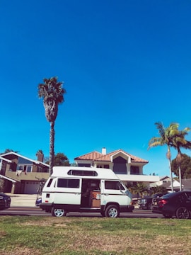 A white camper van is parked on a residential street with its side door open. The background features a modern house with a red-tiled roof and large windows. Tall palm trees rise alongside the house, and the sky above is clear and blue. A few other cars are parked on the street, and there is a well-maintained grassy area in the foreground.