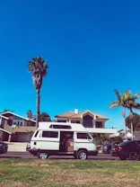 A white camper van is parked on a residential street with its side door open. The background features a modern house with a red-tiled roof and large windows. Tall palm trees rise alongside the house, and the sky above is clear and blue. A few other cars are parked on the street, and there is a well-maintained grassy area in the foreground.