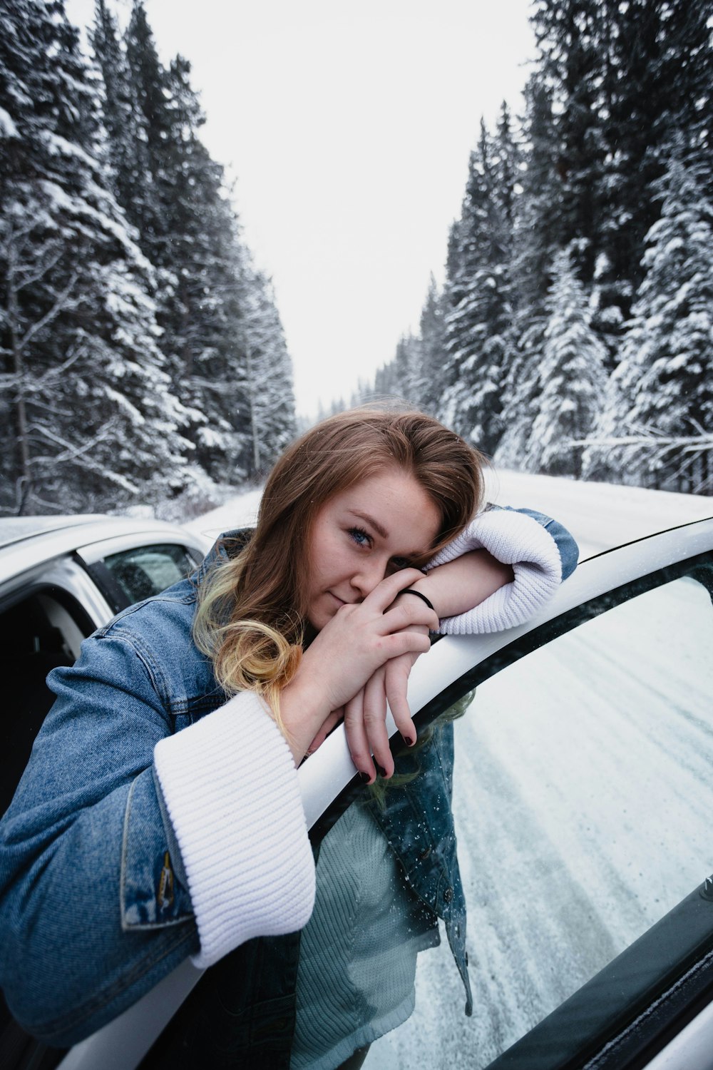 Donna che indossa jeans blu denim appoggiata alla portiera bianca del veicolo sulla strada accanto a pini coperti di neve durante l'inverno