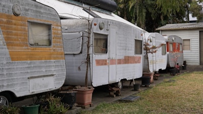 A row of old, weathered caravans are parked closely together on a concrete path. They each show signs of age and wear, with peeling paint and rust spots. Small potted plants and bare trees in containers are placed intermittently in front of the caravans. The surrounding area includes a patchy grass lawn on the right and a background filled with lush green palm trees. A small, plain wooden building with a corrugated metal roof is visible at the end of the line of caravans.