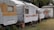 A row of old, weathered caravans are parked closely together on a concrete path. They each show signs of age and wear, with peeling paint and rust spots. Small potted plants and bare trees in containers are placed intermittently in front of the caravans. The surrounding area includes a patchy grass lawn on the right and a background filled with lush green palm trees. A small, plain wooden building with a corrugated metal roof is visible at the end of the line of caravans.