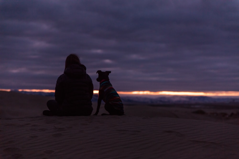 man and dog sitting on sand during sunset
