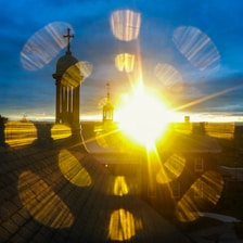 A bright, radiant sunrise peeks over the horizon, illuminating a building with a prominent bell tower topped by a cross. The golden rays of sunlight create dynamic lens flares across the image.