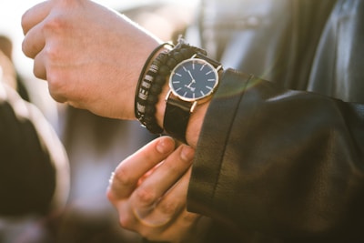 A close-up of a person's wrist adorned with a black watch featuring a minimalist design and a leather strap. The person is also wearing multiple bracelets, including beaded ones, on the same wrist. The outfit includes a black leather jacket, and the lighting creates a warm, soft focus effect.