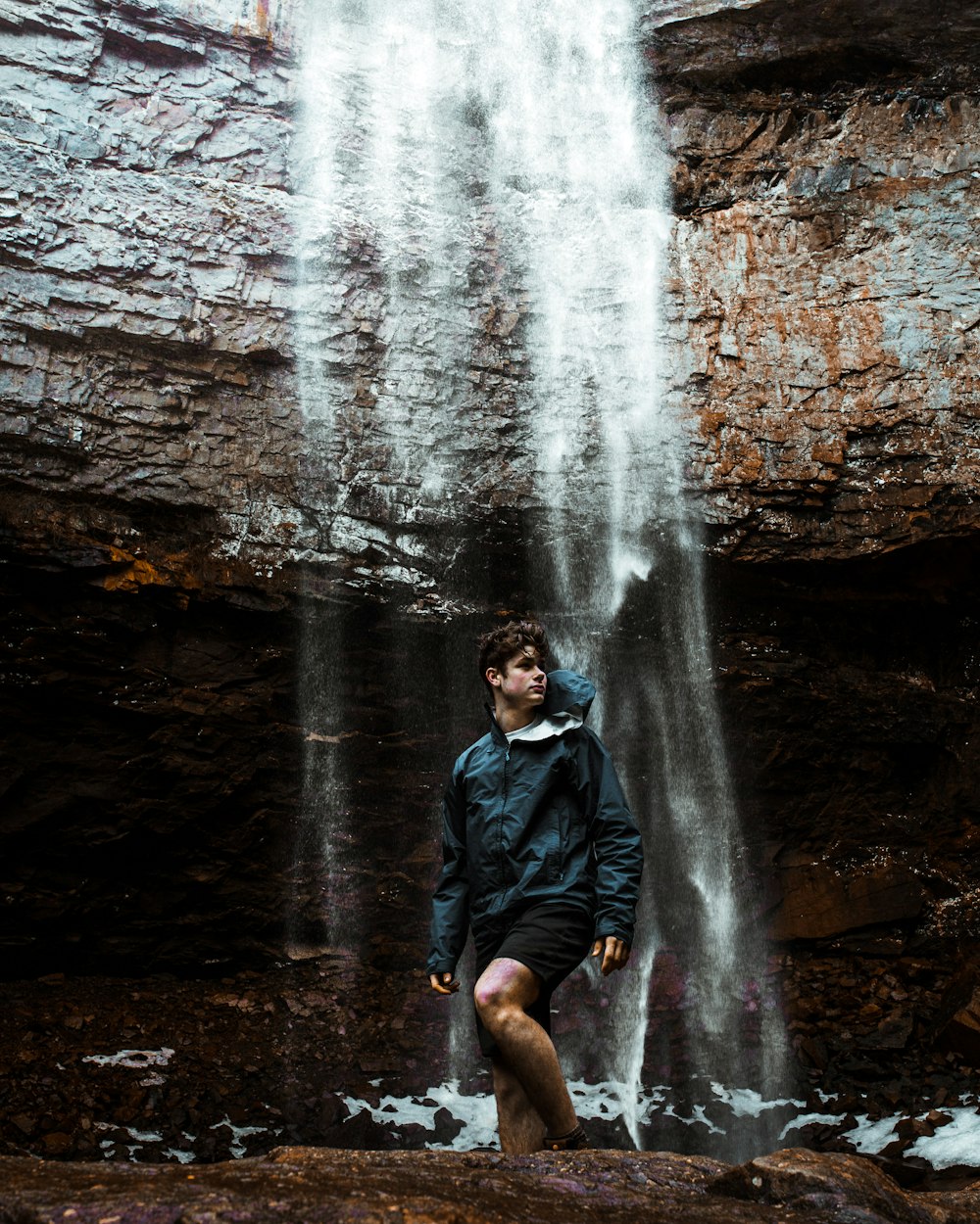 Mann in schwarzer Jacke sitzt auf Felsen vor Wasserfällen