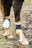 The image features the lower legs of a horse standing on muddy ground. The horse wears protective leg gear, including a fetlock boot and a cannon boot, which are visibly dirty due to the environment. The mud on the ground is mixed with grass, suggesting an outdoor setting.