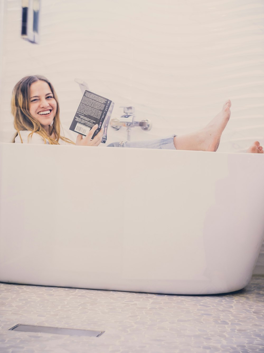 woman in bathtub holding black book while taking selfie