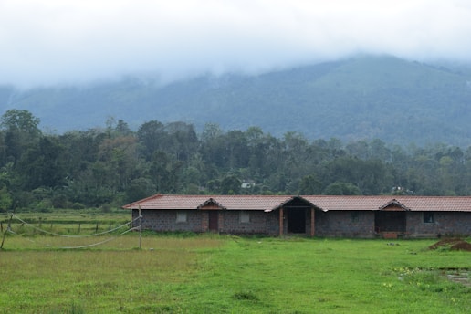 A rural landscape featuring a long, single-story brick building with a red-tiled roof set amidst a large field of green grass. The background is composed of dense forest and a distant mountain partially obscured by low-hanging clouds.