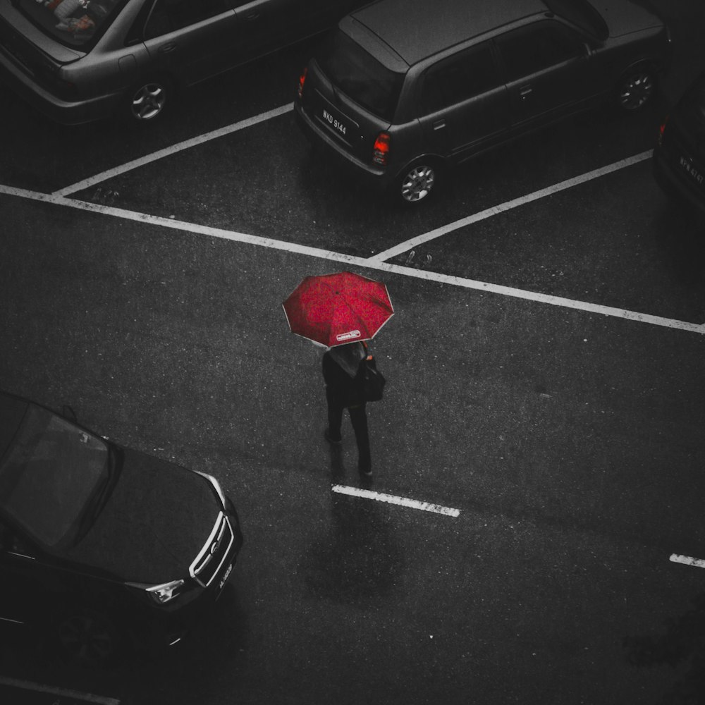 selective color photography of man using red umbrella surrounded by cars