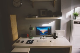 A minimalist and organized desk setup is featured with a contemporary design. The desk has a white surface with a computer monitor displaying a serene nature wallpaper. A green desk lamp is positioned on the left, and a pair of headphones rests next to a closed laptop on the right. Additionally, there is a gaming console with red accents, a smartphone, a notebook, and a keyboard with a mouse arranged neatly. Above the desk, a floating shelf holds a few books, a picture frame, and a small plant. A wall clock hangs on the wall, and soft, natural light filters into the room, enhancing the calming atmosphere.