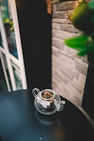 An elegant glass teapot brewing vibrant green tea leaves, surrounded by scattered Indonesian spices on an ivory wooden table.