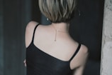 A person with short hair is seen from behind, wearing a black camisole top and a thin chain necklace with a small pendant. The setting appears to be indoors, with dim lighting and a dark background.