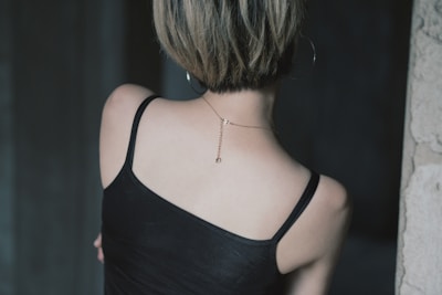 A person with short hair is seen from behind, wearing a black camisole top and a thin chain necklace with a small pendant. The setting appears to be indoors, with dim lighting and a dark background.