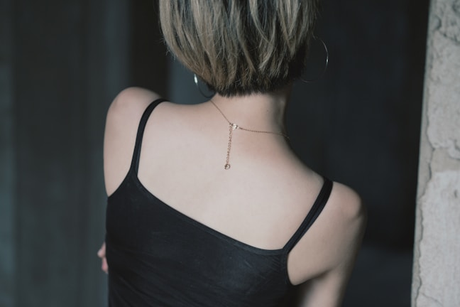 A person with short hair is seen from behind, wearing a black camisole top and a thin chain necklace with a small pendant. The setting appears to be indoors, with dim lighting and a dark background.