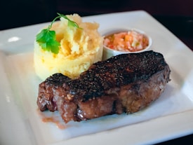 A well-cooked steak is served on a white plate, accompanied by a small portion of mashed potatoes and a side of diced tomato and onion salad. The mashed potatoes are garnished with a parsley leaf.