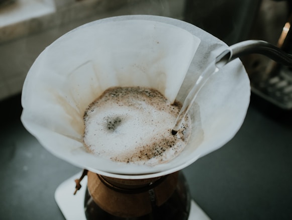 A close-up view of a pour-over coffee brewing process, featuring a white paper filter containing ground coffee with hot water being poured over it from a silver kettle.