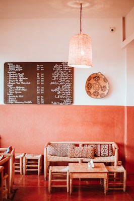 A cozy caf&eacute; interior with warm orange and earthy tones. The space features a wooden table with matching chairs and a couch adorned with patterned cushions. A menu board with text in multiple languages hangs on the wall, alongside a decorative woven basket and a hanging lamp providing soft lighting.
