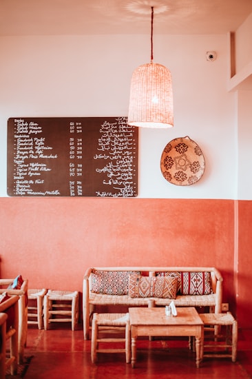 A cozy caf&eacute; interior with warm orange and earthy tones. The space features a wooden table with matching chairs and a couch adorned with patterned cushions. A menu board with text in multiple languages hangs on the wall, alongside a decorative woven basket and a hanging lamp providing soft lighting.