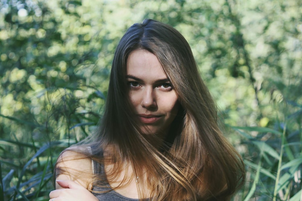 brown haired woman in grey sleeveless top holding her arm standing in front of green leafed trees