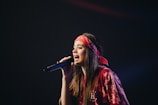 A woman is passionately singing into a microphone. She is wearing a red bandana and a matching red outfit, with long brown hair draped over her shoulders. The background is dark, highlighting her presence on stage.