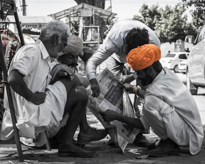Four men are gathered closely together, intently reading a newspaper. They are dressed in traditional attire with one of them wearing a vibrant orange turban that stands out. The scene takes place outdoors on a sunny day, evidenced by the sunlight and shadows cast on the ground. The background includes trees and parked cars, suggesting a roadside or marketplace setting.