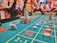 A casino scene with a roulette table features prominently, covered in green felt with colorful numbers and betting squares. Several stacks of poker chips are placed on different numbers, and the hands of several people are visible as they interact with the table. In the background, there are slot machines and more people, creating a vibrant and busy atmosphere.
