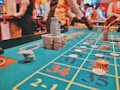 A casino scene with a roulette table features prominently, covered in green felt with colorful numbers and betting squares. Several stacks of poker chips are placed on different numbers, and the hands of several people are visible as they interact with the table. In the background, there are slot machines and more people, creating a vibrant and busy atmosphere.