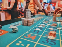 A casino scene with a roulette table features prominently, covered in green felt with colorful numbers and betting squares. Several stacks of poker chips are placed on different numbers, and the hands of several people are visible as they interact with the table. In the background, there are slot machines and more people, creating a vibrant and busy atmosphere.