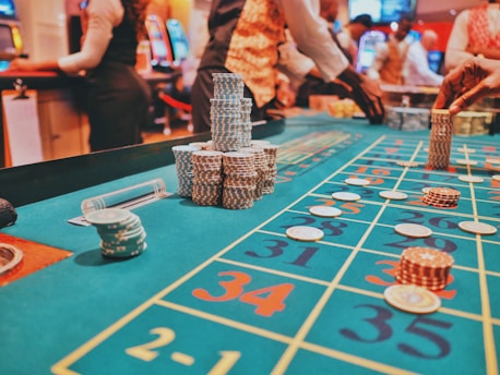 A casino scene with a roulette table features prominently, covered in green felt with colorful numbers and betting squares. Several stacks of poker chips are placed on different numbers, and the hands of several people are visible as they interact with the table. In the background, there are slot machines and more people, creating a vibrant and busy atmosphere.