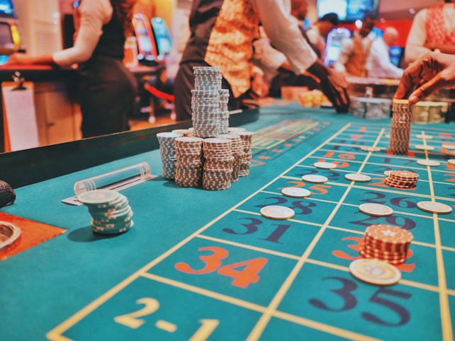 A casino scene with a roulette table features prominently, covered in green felt with colorful numbers and betting squares. Several stacks of poker chips are placed on different numbers, and the hands of several people are visible as they interact with the table. In the background, there are slot machines and more people, creating a vibrant and busy atmosphere.