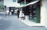 A group of people is gathered outside a shop with a green facade, possibly in a quaint town setting. The store has a striped awning and a large display window showcasing various items. Passersby include a person walking with a child and another older individual further down the street. In the background, a vintage car is parked near the entrance of another building.