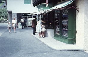 A group of people is gathered outside a shop with a green facade, possibly in a quaint town setting. The store has a striped awning and a large display window showcasing various items. Passersby include a person walking with a child and another older individual further down the street. In the background, a vintage car is parked near the entrance of another building.
