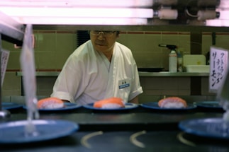 A sushi chef stands behind a counter with plates of sushi on a conveyor belt. The chef is wearing a white uniform and glasses. There are signs with Japanese writing and kitchen equipment in the background.