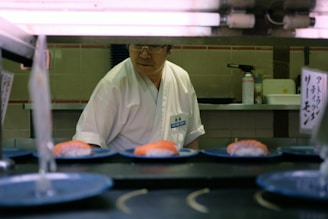 A sushi chef stands behind a counter with plates of sushi on a conveyor belt. The chef is wearing a white uniform and glasses. There are signs with Japanese writing and kitchen equipment in the background.