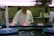 A sushi chef stands behind a counter with plates of sushi on a conveyor belt. The chef is wearing a white uniform and glasses. There are signs with Japanese writing and kitchen equipment in the background.